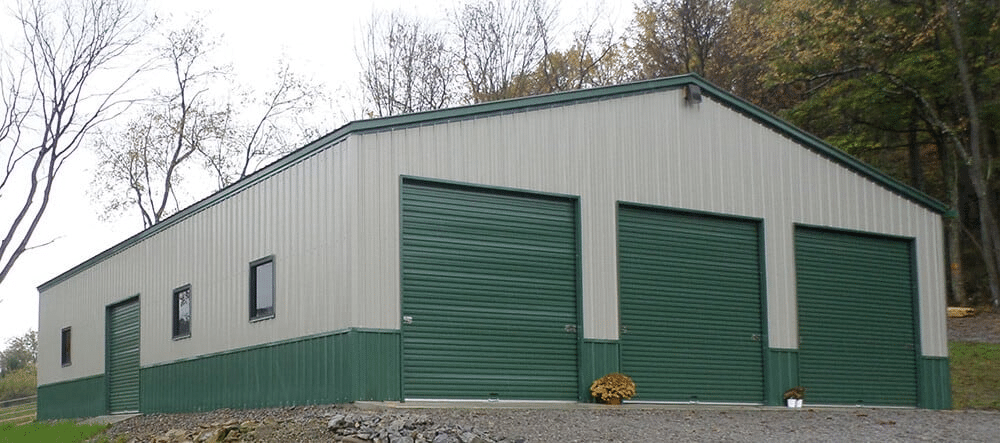 a rectangular steel building in tan and green colors. The building has a sloping roof and 3 large rollup door at the front. There are several windows on each side of the building and a small, rectangular vent on the roof.